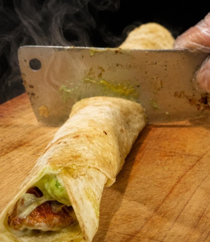 Tortilla de harina enrollada siendo cortada con machete sobre una tabla de madera, con el relleno a la vista